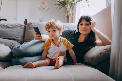 A woman and a young boy are sitting together on a comfortable gray couch in a living room. The woman is leaning back, appearing relaxed, with her hand resting on her head. The boy is sitting in front of her, looking attentively forward as the woman holds a TV remote. A large potted plant and a window with sheer curtains are visible in the background, adding a cozy ambiance to the scene.