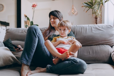 A woman sits comfortably on a couch, holding a young child on her lap. The child looks thoughtful while the woman seems relaxed, holding a smartphone in her hand. The room is decorated with indoor plants and features a modern lamp and wall clock.