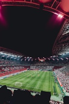 A large, illuminated stadium packed with spectators, showcasing a vibrant soccer match. The field is green with visible players in red and white uniforms, and the stands are filled with red and white-clad fans. The stadium structure is visible above, with bright lights casting a glow.