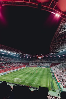 A large, illuminated stadium packed with spectators, showcasing a vibrant soccer match. The field is green with visible players in red and white uniforms, and the stands are filled with red and white-clad fans. The stadium structure is visible above, with bright lights casting a glow.