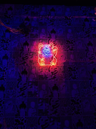 A neon sign of a cat outlined in bright red and blue light is displayed against a backdrop of various dark patterns and illustrations. The vibrant colors of the neon sign create a strong contrast with the darker, monochrome background.