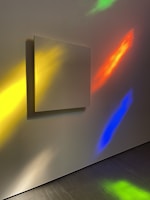 A blank square canvas is mounted on a wall, illuminated by colorful light reflections. The wall features vibrant hues of yellow, red, blue, and green, creating an abstract, artistic atmosphere. The smooth texture of the wall and the shadows cast by the canvas add depth to the scene.
