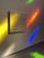 A blank square canvas is mounted on a wall, illuminated by colorful light reflections. The wall features vibrant hues of yellow, red, blue, and green, creating an abstract, artistic atmosphere. The smooth texture of the wall and the shadows cast by the canvas add depth to the scene.