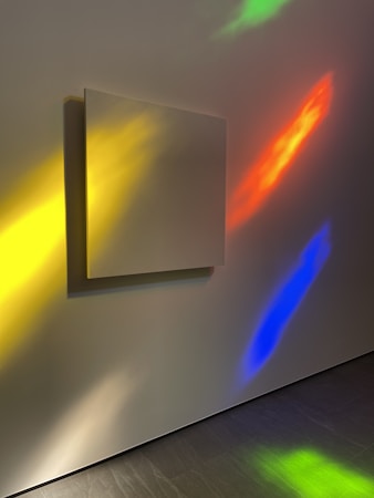 A blank square canvas is mounted on a wall, illuminated by colorful light reflections. The wall features vibrant hues of yellow, red, blue, and green, creating an abstract, artistic atmosphere. The smooth texture of the wall and the shadows cast by the canvas add depth to the scene.
