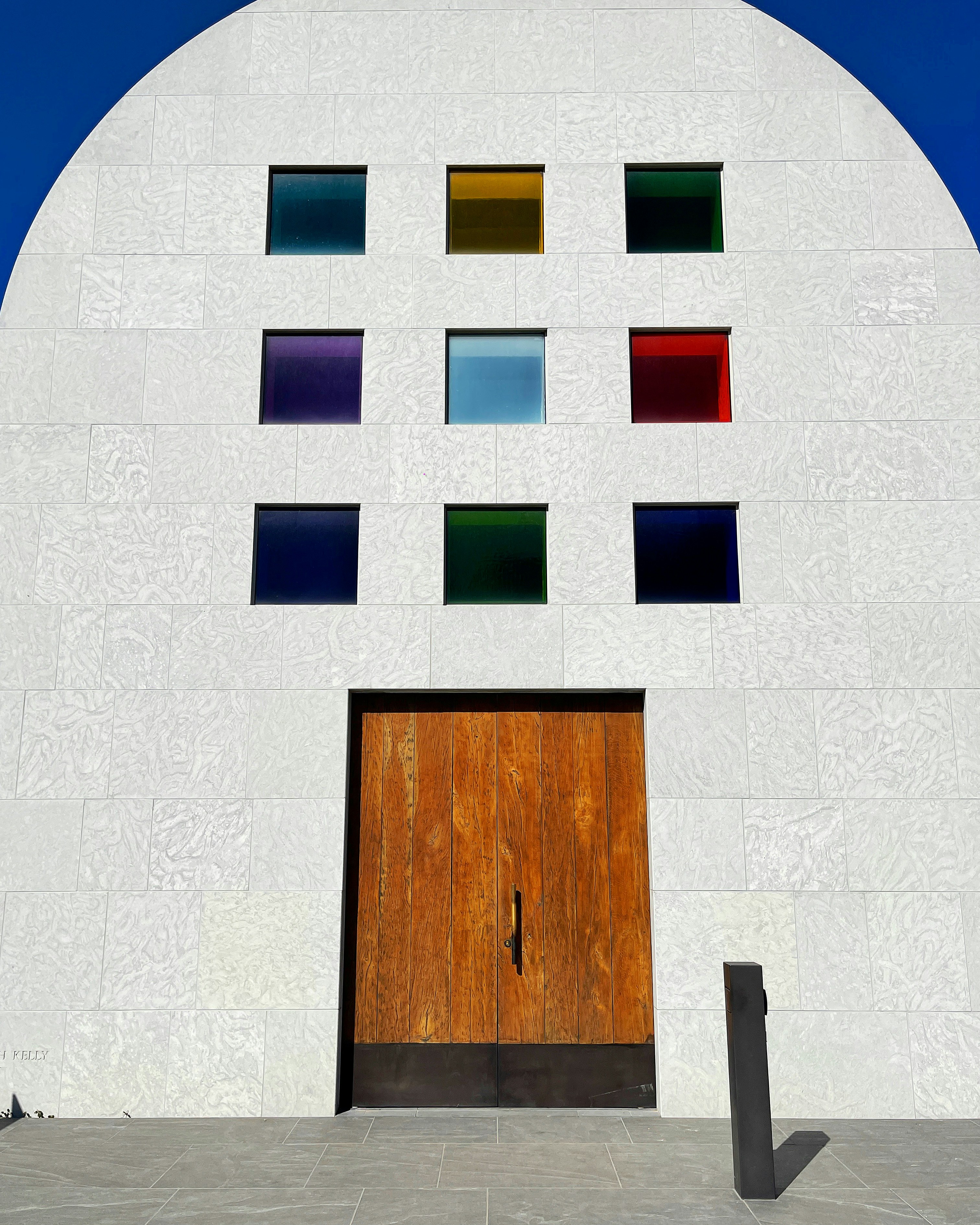 A modern building facade with a large wooden door surrounded by white textured tiles. Nine square windows, each with a different color, are arranged above the door. The colors of the windows are diverse and vibrant, including shades of blue, red, green, and purple.