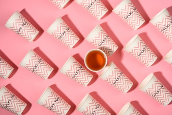 Several paper cups with a zigzag pattern and the words 'Lucky You' are arranged on a pink background. One of the cups is filled with a brown liquid.