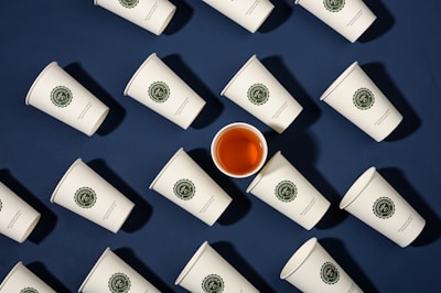 A series of white paper cups with green logos arranged on a dark blue background. Among them, one cup is filled with a dark orange liquid, possibly tea, standing out from the rest.