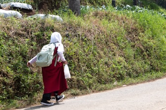 A person wearing a white hijab and a red uniform is walking along a road beside a grassy embankment. The person is carrying a large white backpack and holding a plastic bag in their hand. The setting appears to be rural, with greenery and vegetation surrounding the pathway.