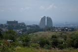 A cityscape featuring a pair of modern skyscrapers with a unique curved design. Surrounded by residential buildings and lush greenery in the foreground, the scene includes solar panels and a hilly landscape. The sky is overcast, adding a serene mood to the image.