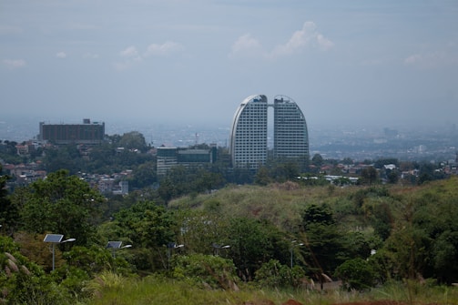 A cityscape featuring a pair of modern skyscrapers with a unique curved design. Surrounded by residential buildings and lush greenery in the foreground, the scene includes solar panels and a hilly landscape. The sky is overcast, adding a serene mood to the image.
