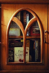 A wooden cabinet with an arched glass door reveals several books stacked inside. Prominent titles include works by Paul Coelho and National Geographic. The warm wood tones of the cabinet contribute to a cozy and inviting ambiance.