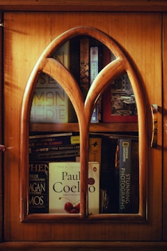 A wooden cabinet with an arched glass door reveals several books stacked inside. Prominent titles include works by Paul Coelho and National Geographic. The warm wood tones of the cabinet contribute to a cozy and inviting ambiance.