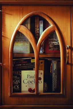 A wooden cabinet with an arched glass door reveals several books stacked inside. Prominent titles include works by Paul Coelho and National Geographic. The warm wood tones of the cabinet contribute to a cozy and inviting ambiance.