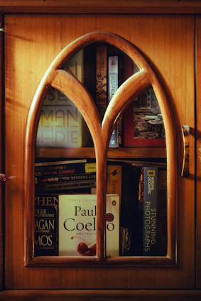 A wooden cabinet with an arched glass door reveals several books stacked inside. Prominent titles include works by Paul Coelho and National Geographic. The warm wood tones of the cabinet contribute to a cozy and inviting ambiance.