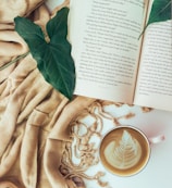 An open book with visible text is placed on a soft beige fabric alongside two large green leaves resting on the pages. A cup of cappuccino with latte art depicting a leaf pattern sits nearby, adding an artistic touch to the cozy arrangement.