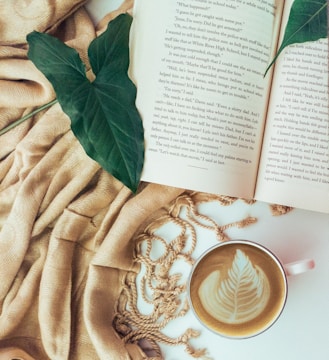 An open book with visible text is placed on a soft beige fabric alongside two large green leaves resting on the pages. A cup of cappuccino with latte art depicting a leaf pattern sits nearby, adding an artistic touch to the cozy arrangement.