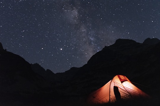 A brightly illuminated tent is set up against a backdrop of a starry night sky, featuring the Milky Way. Silhouettes of mountains can be seen in the distance.