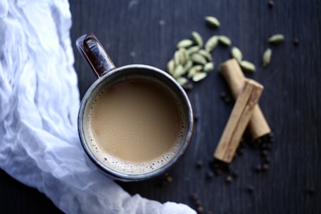 A top view of a ceramic mug filled with creamy tea, placed on a dark wooden surface. Surrounding the mug are whole cardamom pods, cinnamon sticks, and a white cloth on the left side. The arrangement has a cozy and inviting feel.