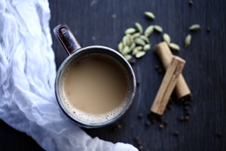 A top view of a ceramic mug filled with creamy tea, placed on a dark wooden surface. Surrounding the mug are whole cardamom pods, cinnamon sticks, and a white cloth on the left side. The arrangement has a cozy and inviting feel.