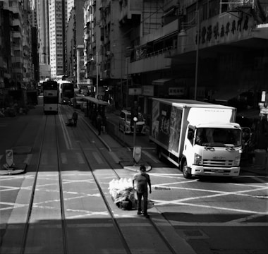 An urban street scene featuring tall buildings lining both sides of the road. A tram approaches in the distance, while a truck is parked on the right side of the road. A man, wearing a mask, walks across the street pulling a cart filled with stacked bags. The buildings and streets are bustling, creating a sense of urban life.