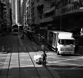 An urban street scene featuring tall buildings lining both sides of the road. A tram approaches in the distance, while a truck is parked on the right side of the road. A man, wearing a mask, walks across the street pulling a cart filled with stacked bags. The buildings and streets are bustling, creating a sense of urban life.