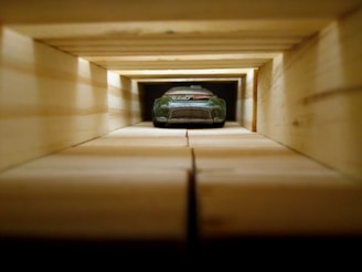 A toy car is placed at the end of a long, tunnel-like structure made of wooden blocks. The perspective creates a sense of depth, with the wooden blocks aligned in a grid pattern leading to the car.