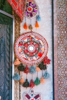 An intricate, colorful wall hanging featuring a circular design with various geometric patterns and tassels. The piece is adorned with vibrant colors such as red, blue, and orange, and is surrounded by a detailed carpet with floral motifs.