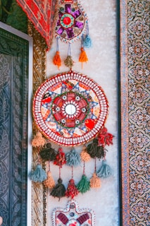 An intricate, colorful wall hanging featuring a circular design with various geometric patterns and tassels. The piece is adorned with vibrant colors such as red, blue, and orange, and is surrounded by a detailed carpet with floral motifs.