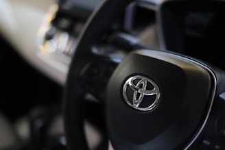 A close-up view of a car's steering wheel with a prominent Toyota logo in the center. The background is slightly blurred, focusing attention on the steering wheel and logo.