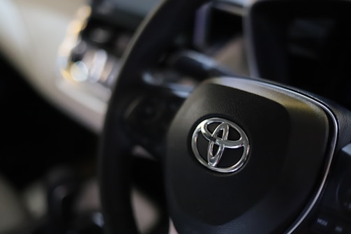 A close-up view of a car's steering wheel with a prominent Toyota logo in the center. The background is slightly blurred, focusing attention on the steering wheel and logo.
