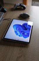 A tablet displaying a vibrant blue abstract design is placed on a wooden table. A computer mouse and a game controller are also on the table, while the edge of a laptop is visible.