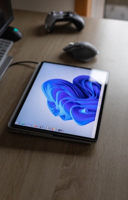 A tablet displaying a vibrant blue abstract design is placed on a wooden table. A computer mouse and a game controller are also on the table, while the edge of a laptop is visible.