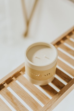 A beige-colored candle in a jar sits on a wooden slatted surface, suggesting a minimalist and cozy setting. The label on the candle reads 'Nomad' and features a subtle design.