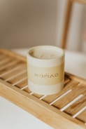 A minimalist cylindrical candle labeled 'NOMAD' is placed on a wooden slat tray. The setting appears calm and aesthetic with a focus on the simple yet elegant design of the candle. The lighting is soft, creating a warm ambiance.