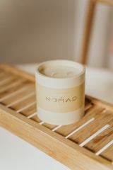 A minimalist cylindrical candle labeled 'NOMAD' is placed on a wooden slat tray. The setting appears calm and aesthetic with a focus on the simple yet elegant design of the candle. The lighting is soft, creating a warm ambiance.