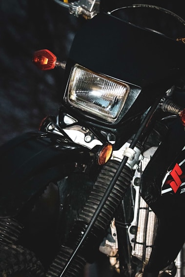 The image depicts a close-up of a motorcycle showing its headlight, indicators, and part of the front suspension system. The design features include a square headlight encased in a black frame, complemented by orange indicators on the sides. The brand logo is visible on the side panel, and the front fork and suspension components are highlighted.