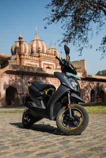 A sleek, modern scooter is parked on a cobblestone path in front of a historical building with domes and intricate architecture. The sun casts clear shadows, indicating a bright day.