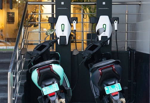 Two electric scooters are parked and connected to a charging station. The station is mounted against a railing, and one of the scooters is mint green while the other is black. There are license plates on both scooters, and the background shows an indoor area with plants and warm lighting.