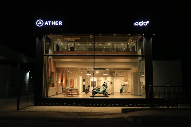 A modern showroom with large glass windows illuminated at night. Inside, there are two electric scooters displayed under warm lighting, alongside sleek wooden furniture and minimalist decor. The showroom is well-organized, and the lighting highlights the clean, contemporary design.