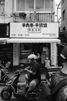 A bustling street scene featuring people riding scooters in front of a building with signage in Chinese characters. The building's facade includes a balcony with clothes hanging, and pedestrians can be seen near the entrance.