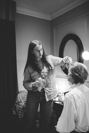 A woman in a denim shirt is doing another person's hair in a room with a mirror and a lit bulb. She holds a can, possibly hairspray, while focusing intently on styling. The person sitting has their back to the camera, wearing a white shirt.