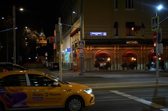 A street scene at night featuring a yellow taxi with Cyrillic writing on its side, parked near a building named 'MICHEL.' The building has multiple arched windows with warm lighting emanating from within, suggesting a cozy interior. There are street signs in Cyrillic, street lamps, and a distant, illuminated tower visible in the background.