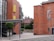 A modern architectural building with a mix of brick and glass elements. The brick portion features multiple small windows and a large sign displaying the word 'ADAMS'. The glass section of the building includes large windows with yellow accents. A small tree and lamp posts are present in the foreground, and another brick building with a traditional design is partially visible in the background.