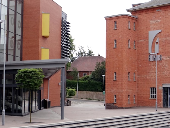 A modern architectural building with a mix of brick and glass elements. The brick portion features multiple small windows and a large sign displaying the word 'ADAMS'. The glass section of the building includes large windows with yellow accents. A small tree and lamp posts are present in the foreground, and another brick building with a traditional design is partially visible in the background.