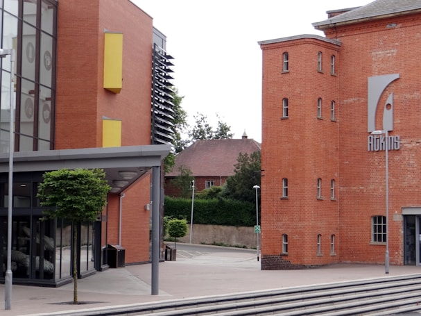 A modern architectural building with a mix of brick and glass elements. The brick portion features multiple small windows and a large sign displaying the word 'ADAMS'. The glass section of the building includes large windows with yellow accents. A small tree and lamp posts are present in the foreground, and another brick building with a traditional design is partially visible in the background.