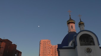 A church with blue domes and golden crosses is set against a clear sky. A half-moon is visible next to a multi-story residential building, and the structures are bathed in the warm light of the setting or rising sun.