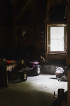 A dimly lit garage or workshop with a window allowing minimal natural light. A purple lawnmower or small tractor sits in the middle, surrounded by various tools and equipment. The walls and floor are wooden and concrete, enhancing the rustic and slightly neglected ambiance.