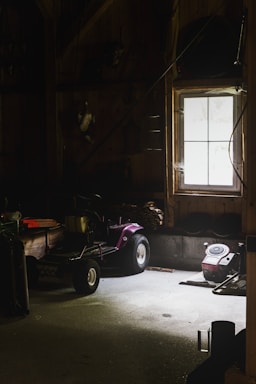 A dimly lit garage or workshop with a window allowing minimal natural light. A purple lawnmower or small tractor sits in the middle, surrounded by various tools and equipment. The walls and floor are wooden and concrete, enhancing the rustic and slightly neglected ambiance.