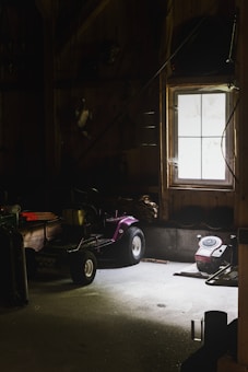 A dimly lit garage or workshop with a window allowing minimal natural light. A purple lawnmower or small tractor sits in the middle, surrounded by various tools and equipment. The walls and floor are wooden and concrete, enhancing the rustic and slightly neglected ambiance.