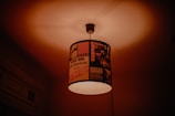 A ceiling light with a lampshade decorated with vintage rock band posters and album covers. The ambient light creates a warm glow, casting soft shadows on the ceiling.